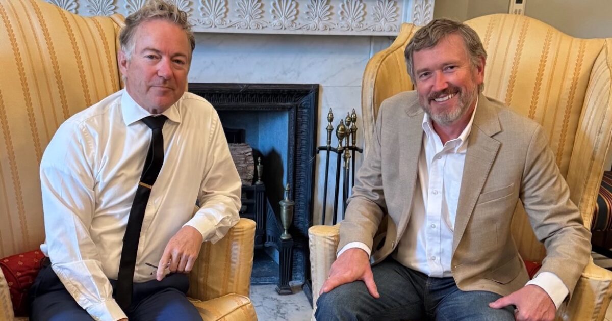 Two men sitting in armchairs, one in a white shirt and tie, the other in a beige blazer, engaged in conversation in a cozy room with a fireplace.