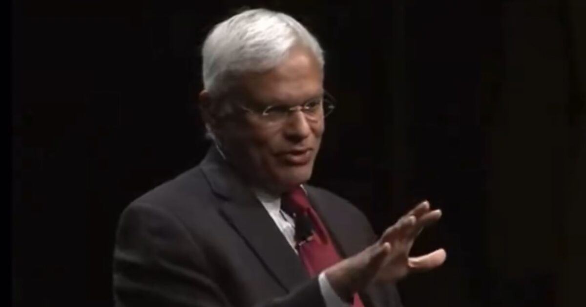 Business leader delivering a presentation, gesturing with hand, wearing glasses and a suit, against a dark background.