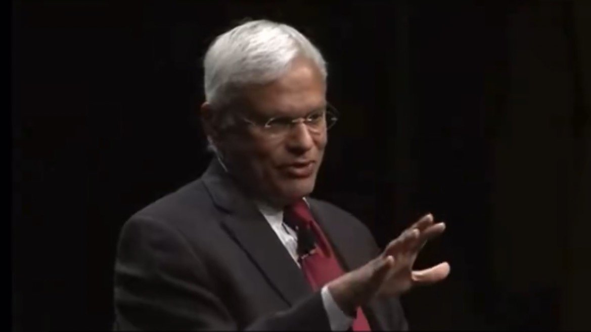 Business leader delivering a presentation, gesturing with hand, wearing glasses and a suit, against a dark background.