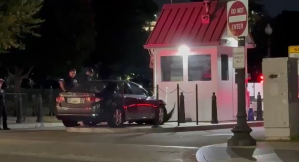 Police officers interact with a vehicle near a security booth at night, highlighting security measures in urban areas.