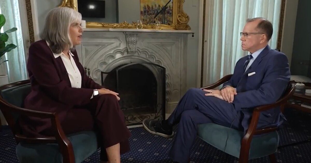 A woman in a burgundy suit and a man in a blue suit engage in a serious conversation in a formal setting with a fireplace in the background.