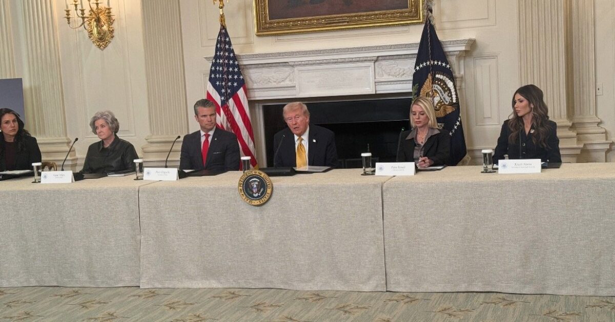 Donald Trump speaks during a roundtable discussion with advisors in the White House, featuring flags and an official presidential seal.