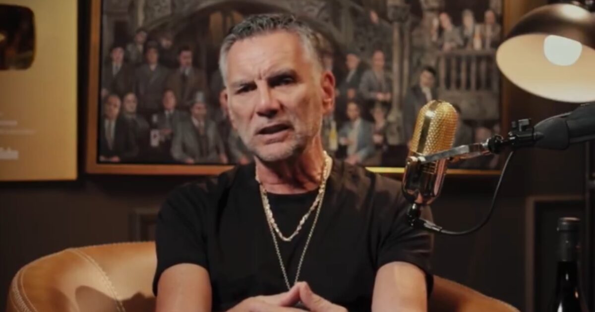 Man in a black shirt and gold chain sits in front of a vintage microphone, discussing topics in a cozy studio setting with framed portraits in the background.
