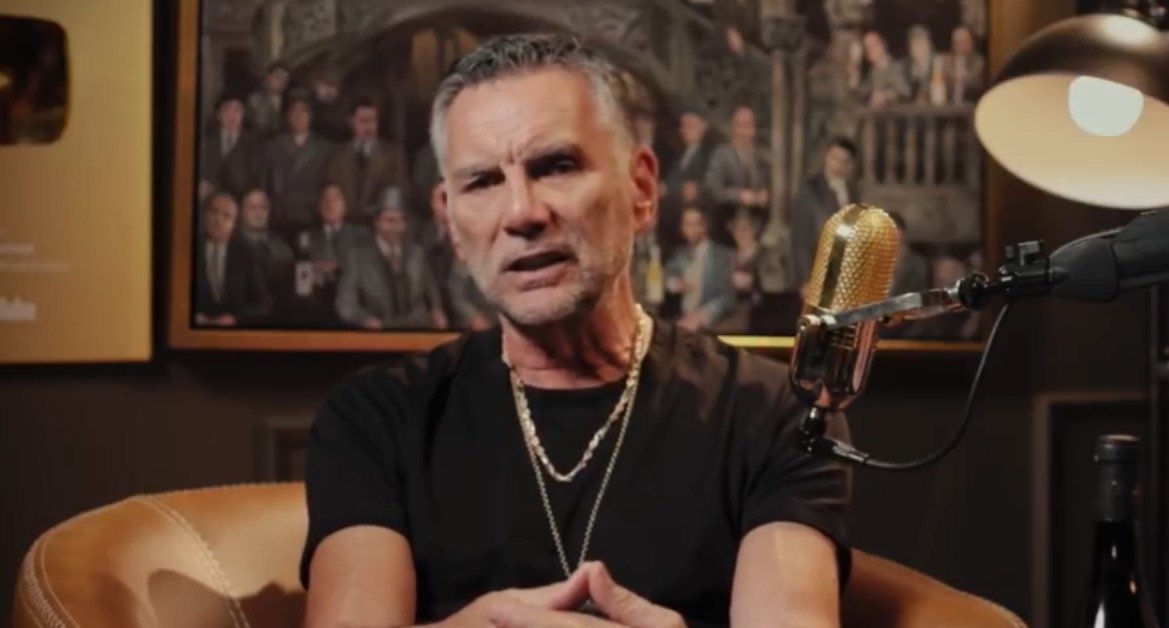 Man in a black shirt and gold chain sits in front of a vintage microphone, discussing topics in a cozy studio setting with framed portraits in the background.