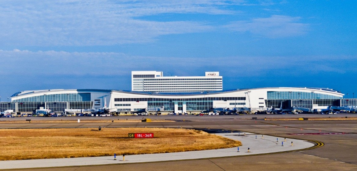 Modern airport terminal with multiple gates, aircraft parked on the tarmac, and a hotel in the background under a clear blue sky.