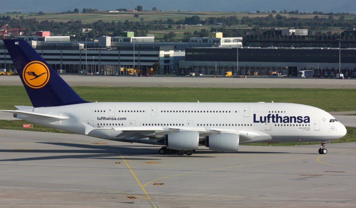 Lufthansa A380 aircraft taxiing on the runway, showcasing its distinctive livery and large size against a backdrop of an airport terminal.