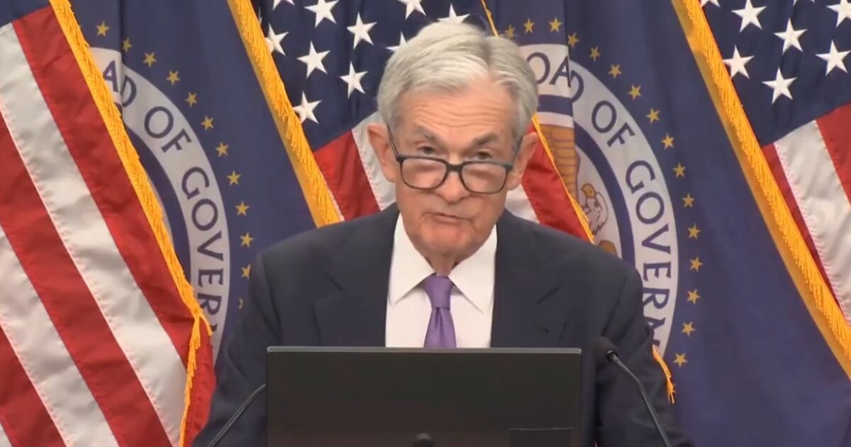 Jerome Powell delivers a speech at a press conference, with U.S. flags and the Federal Reserve seal in the background.
