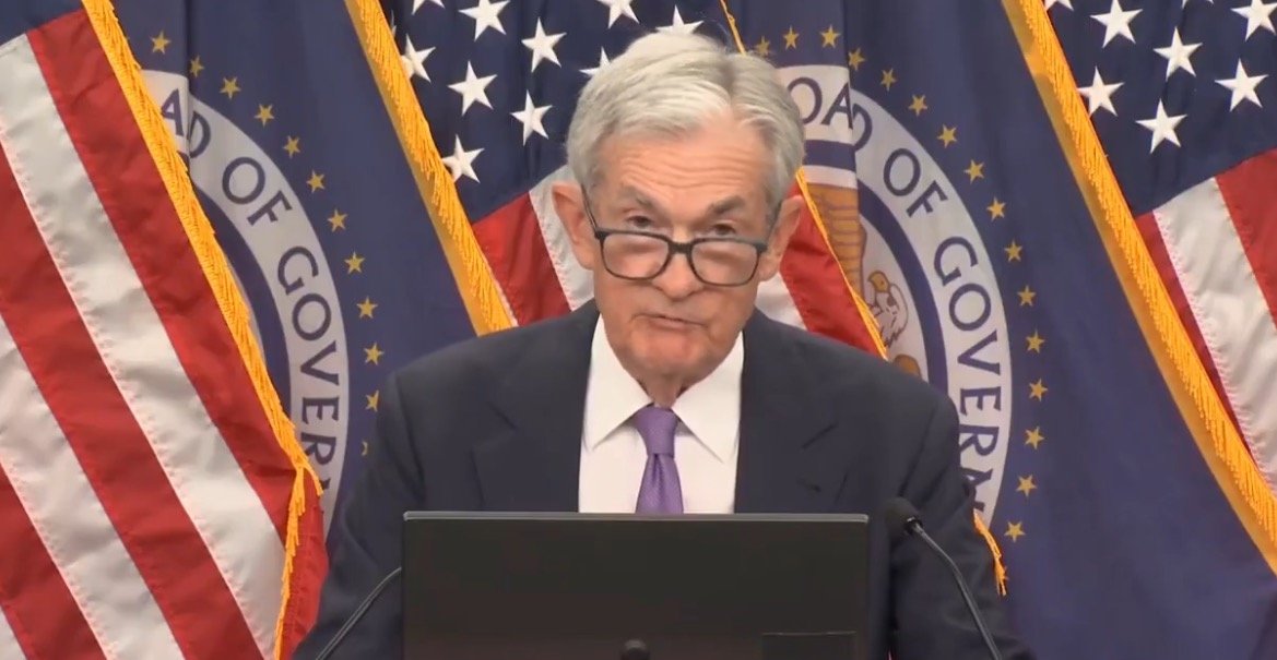 Jerome Powell delivers a speech at a press conference, with U.S. flags and the Federal Reserve seal in the background.