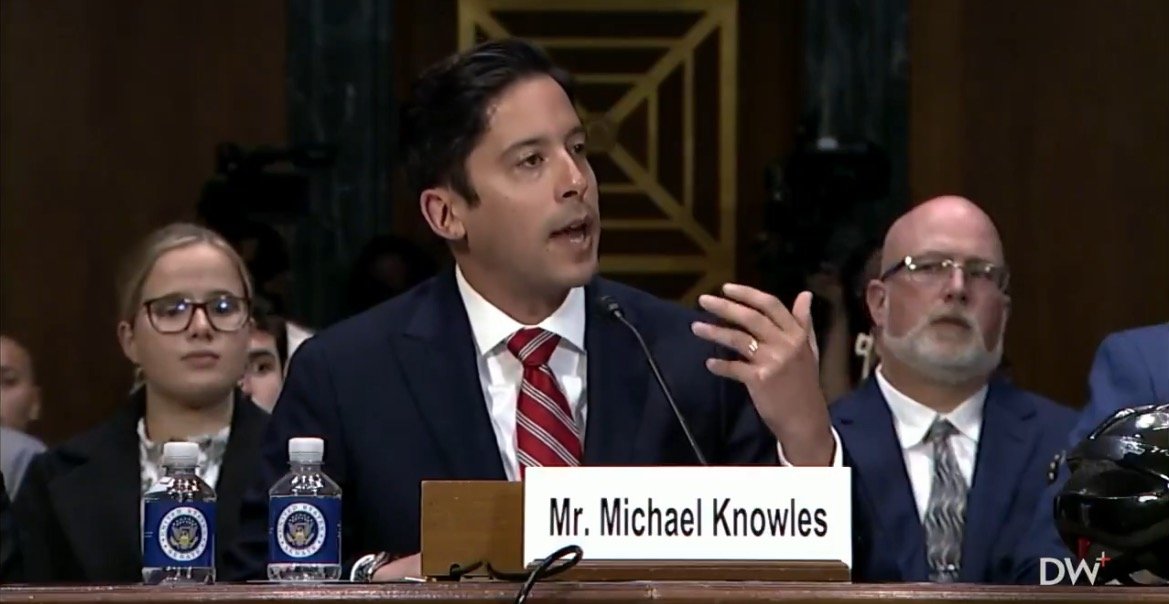 Michael Knowles testifies at a Senate hearing, discussing key issues while seated alongside other attendees, with water bottles and a nameplate visible.