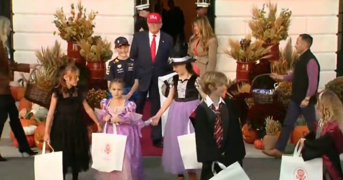 Children in Halloween costumes participate in a festive event at the White House, accompanied by adults and surrounded by autumn decorations.