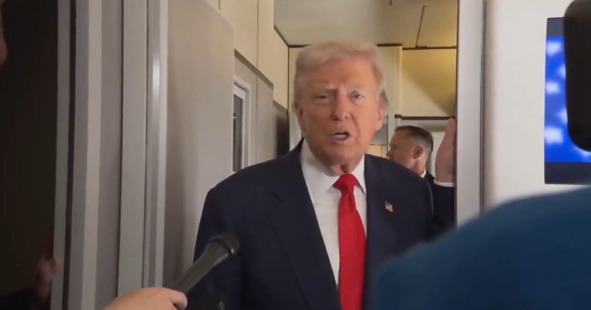 Donald Trump speaking to reporters in a corridor, wearing a dark suit and red tie, while a microphone is pointed towards him.