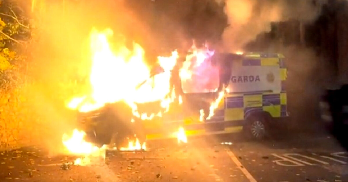 A Garda van engulfed in flames, emitting thick smoke and intense fire, highlighting a scene of emergency and chaos.