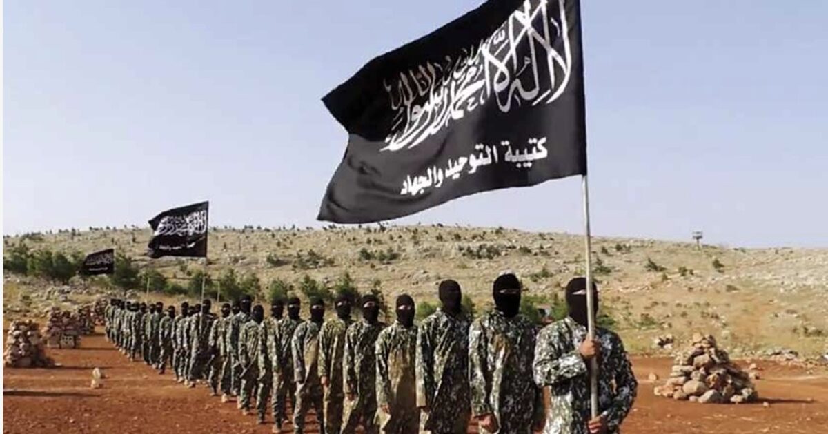Militant group members in camouflage uniforms participate in a formation display while holding black flags in a rural setting.