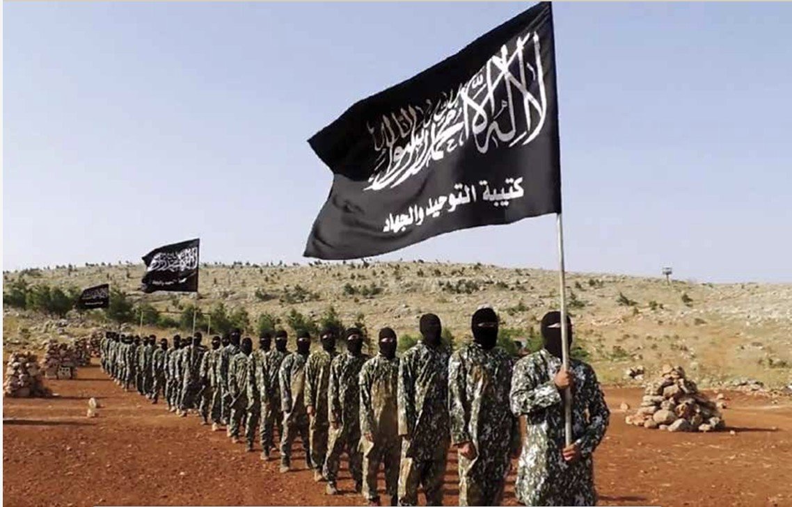 Militant group members in camouflage uniforms participate in a formation display while holding black flags in a rural setting.