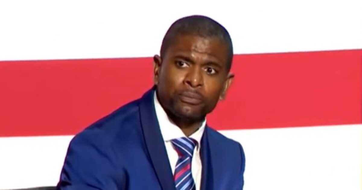 Man in a blue suit with a concerned expression sits against a backdrop of red and white stripes, conveying a serious moment during a discussion or event.
