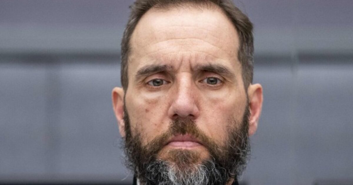 Serious-looking man with a beard wearing a black and purple outfit, standing in a modern indoor setting.