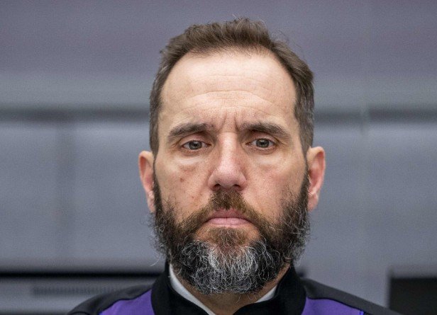 Serious-looking man with a beard wearing a black and purple outfit, standing in a modern indoor setting.