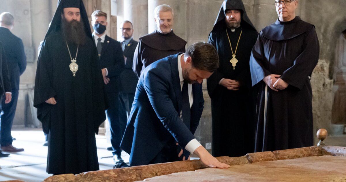 A group of clergy members observes a man in a suit touching a historic stone slab in a religious setting.