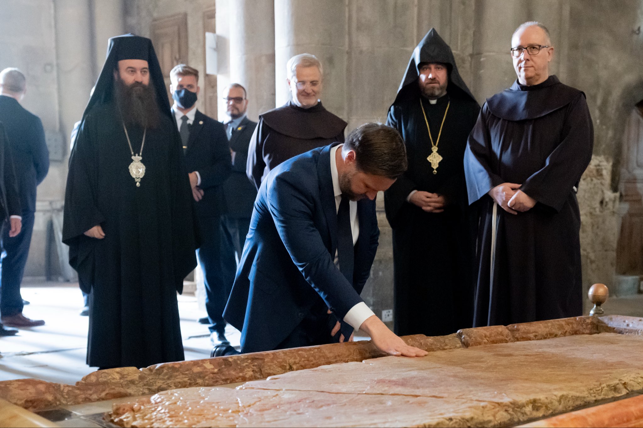A group of clergy members observes a man in a suit touching a historic stone slab in a religious setting.