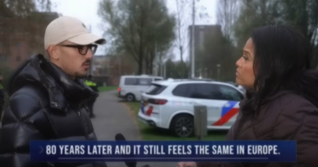 Interview between two individuals discussing historical events and their impact in Europe, with police vehicles in the background.