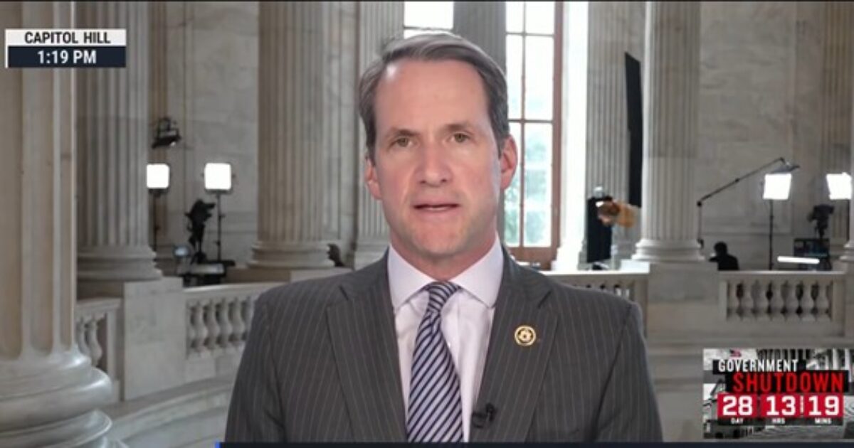 Man in a suit speaking from Capitol Hill, with a backdrop of marble columns and media equipment, discussing government shutdown.