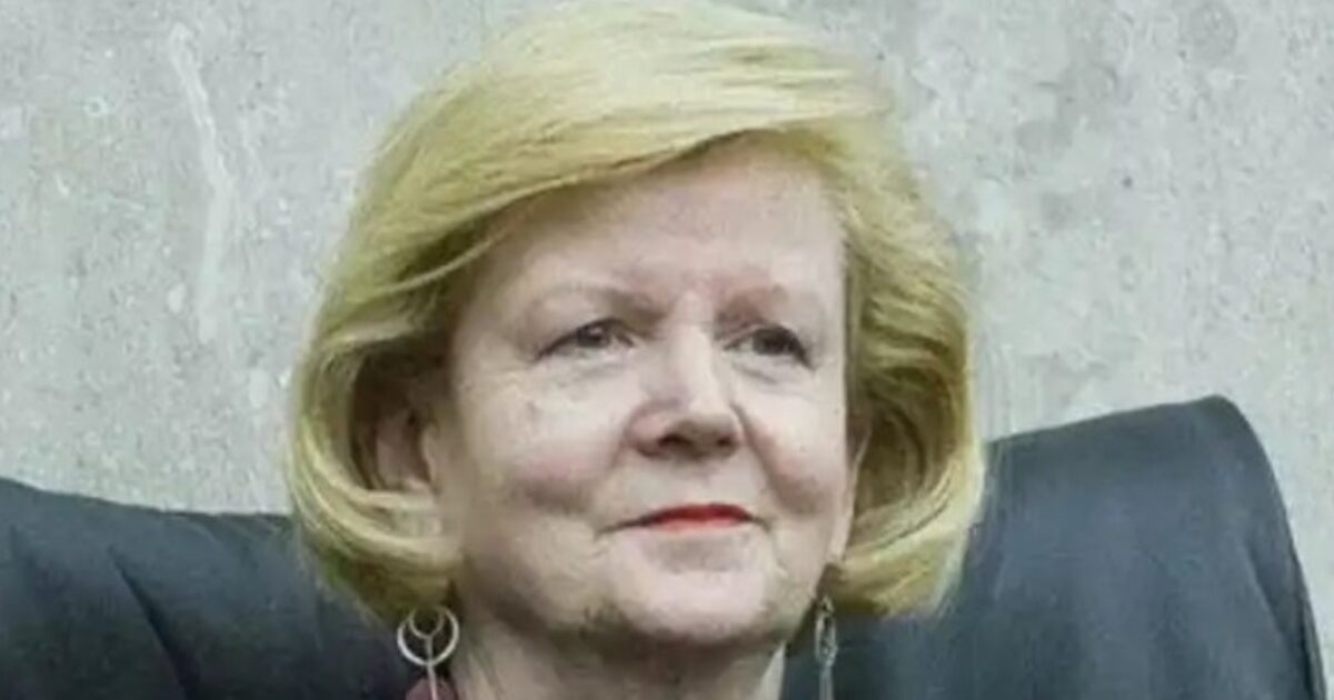 Portrait of a woman with blonde hair and earrings, seated in a formal setting, exuding a confident demeanor.