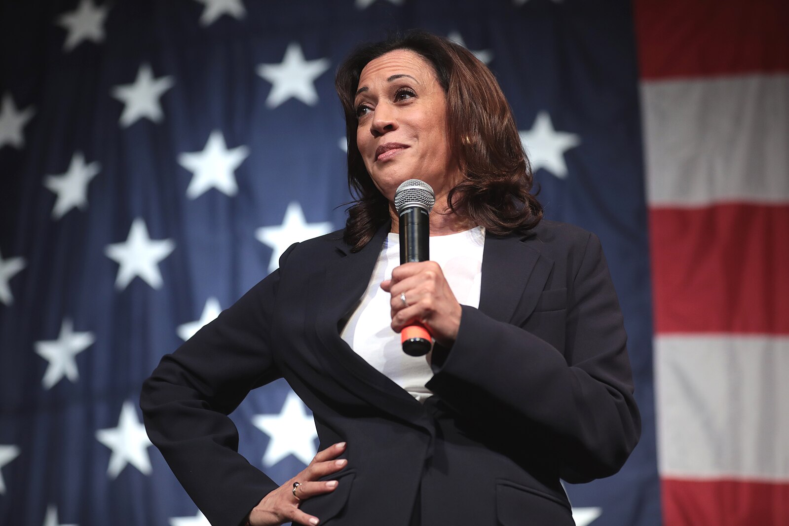 Kamala Harris speaking at a podium with a microphone, set against a backdrop of the American flag.
