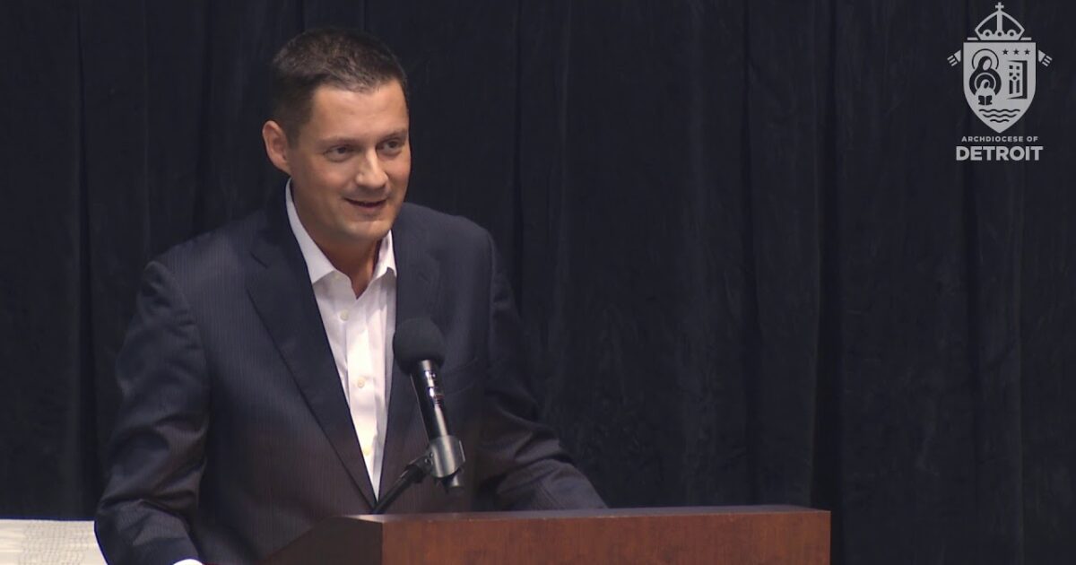 A speaker in a suit addressing an audience at an event hosted by the Archdiocese of Detroit.