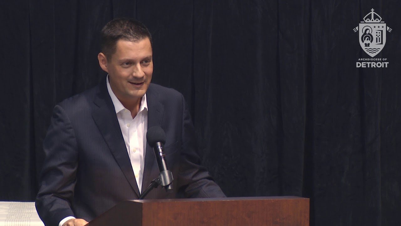 A speaker in a suit addressing an audience at an event hosted by the Archdiocese of Detroit.