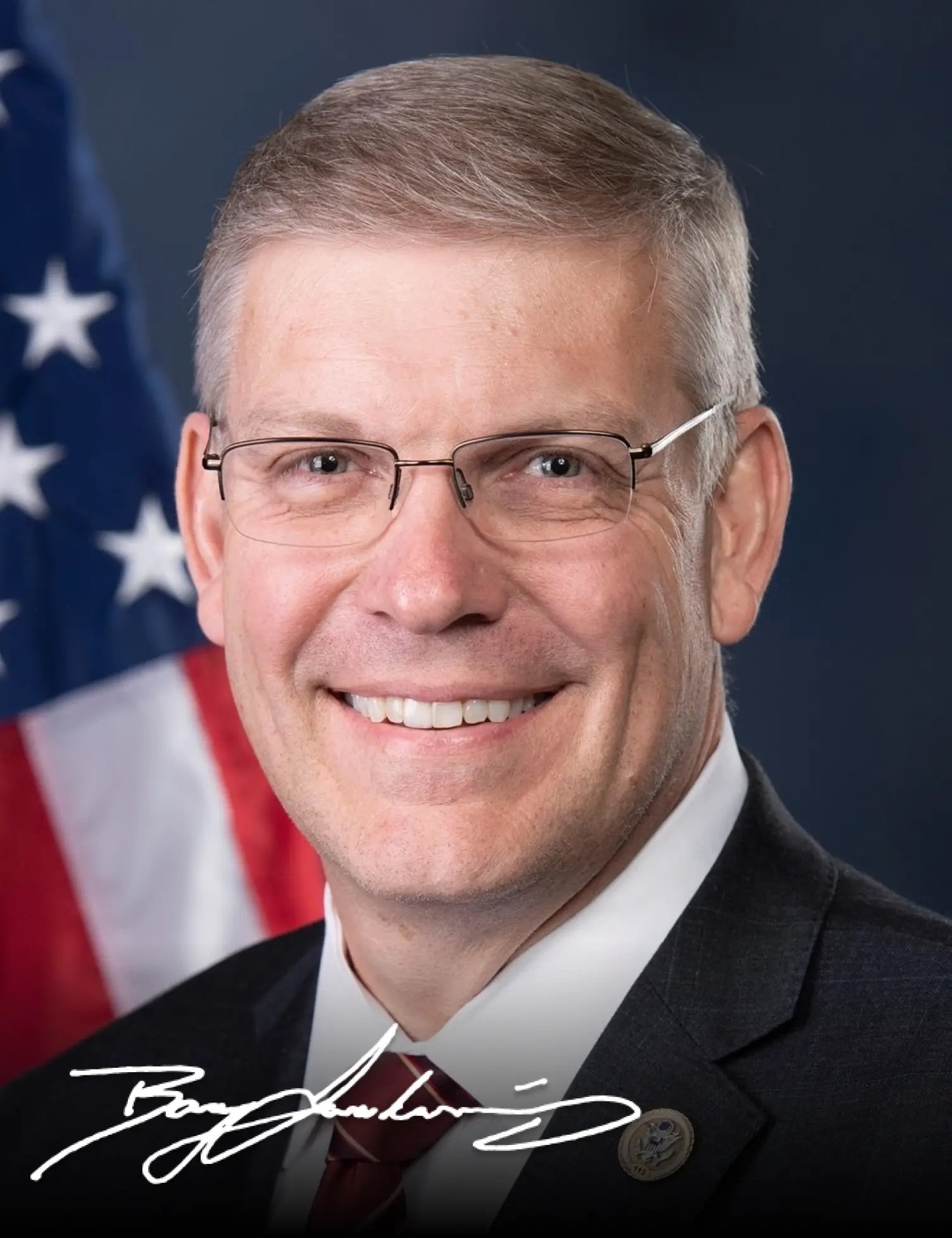 Portrait of a smiling man in a suit with glasses, standing in front of an American flag.