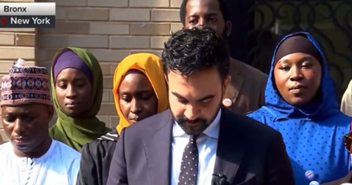 A diverse group of individuals stands together in the Bronx, New York, with a man in a suit reading from a document.