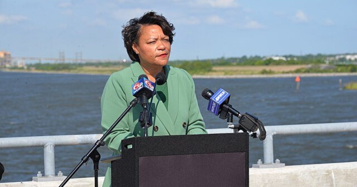 A woman in a green blazer speaks at a podium by a river, with microphones and a scenic background under a clear blue sky.