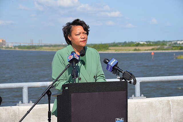 A woman in a green blazer speaks at a podium by a river, with microphones and a scenic background under a clear blue sky.