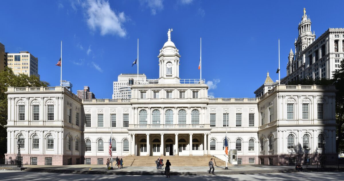 New York City Hall, a historic government building, features classic architecture and is surrounded by urban greenery and modern skyscrapers under a clear blue sky.
