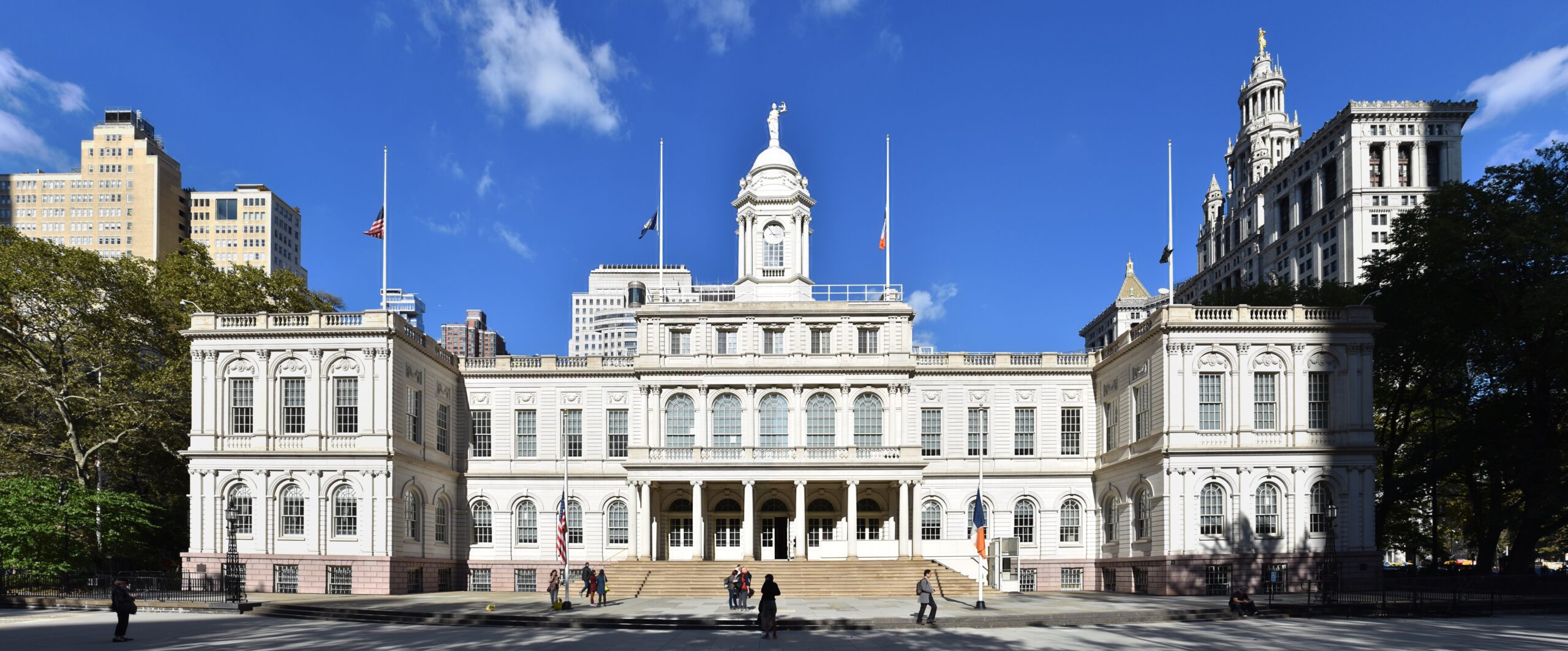 New York City Hall, a historic government building, features classic architecture and is surrounded by urban greenery and modern skyscrapers under a clear blue sky.