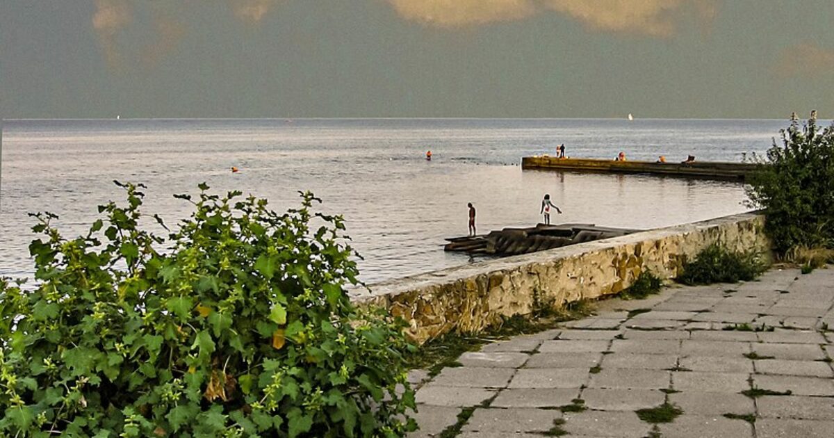 Scenic waterfront view with people enjoying the water, lush greenery, and a cloudy sky reflecting on the calm sea.