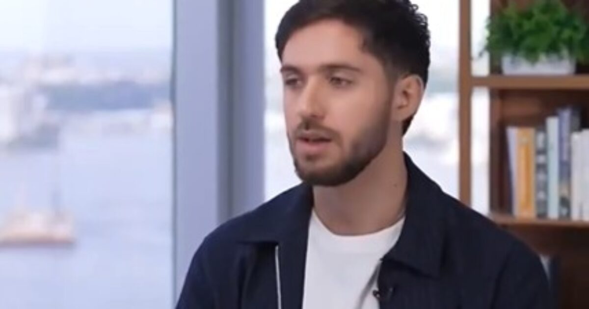 Young man with a beard speaking in an interview setting, with a cityscape visible in the background and a bookshelf beside him.