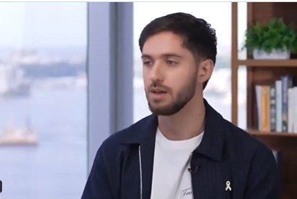 Young man with a beard speaking in an interview setting, with a cityscape visible in the background and a bookshelf beside him.