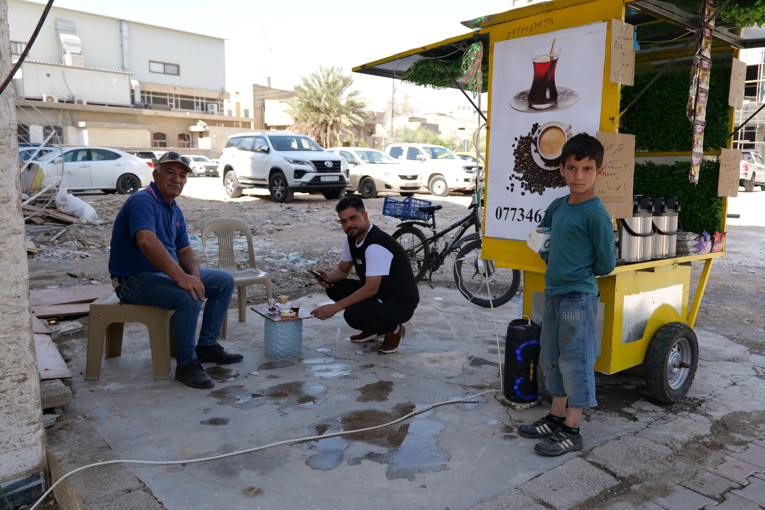 Two men and a boy gather around a small coffee cart in a busy outdoor setting, showcasing a casual community atmosphere.