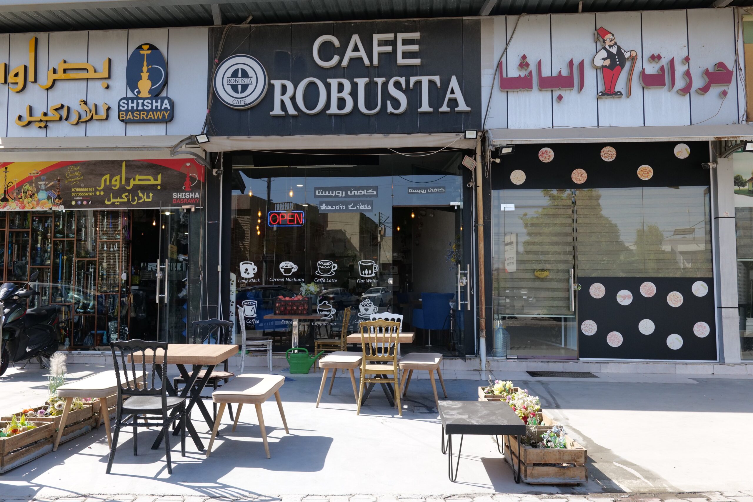 Exterior view of Cafe Robusta featuring outdoor seating, an open sign, and nearby shisha shop, showcasing a vibrant atmosphere for coffee and socializing.