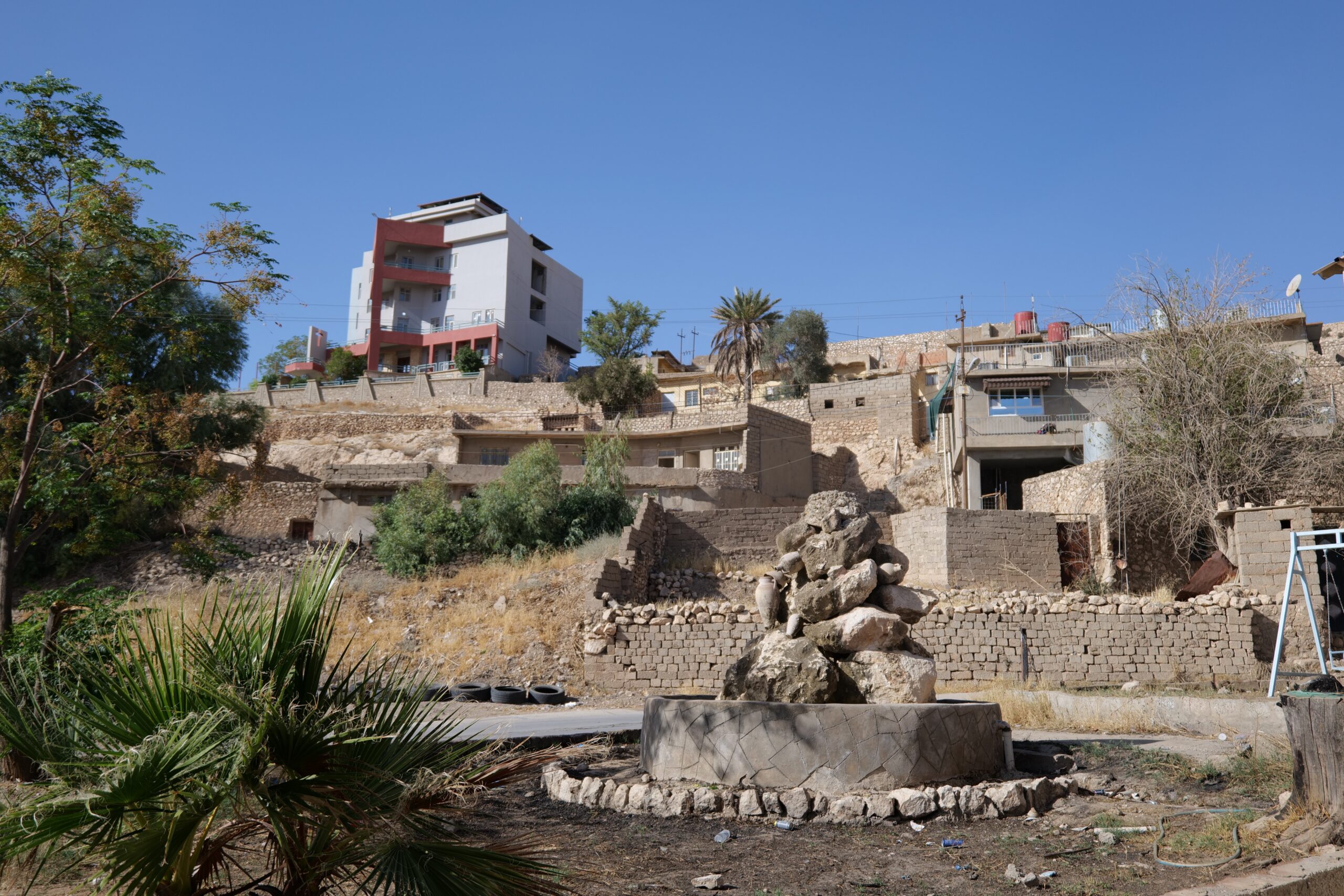 View of a hillside neighborhood featuring a modern building, traditional stone houses, and a stone fountain surrounded by vegetation.