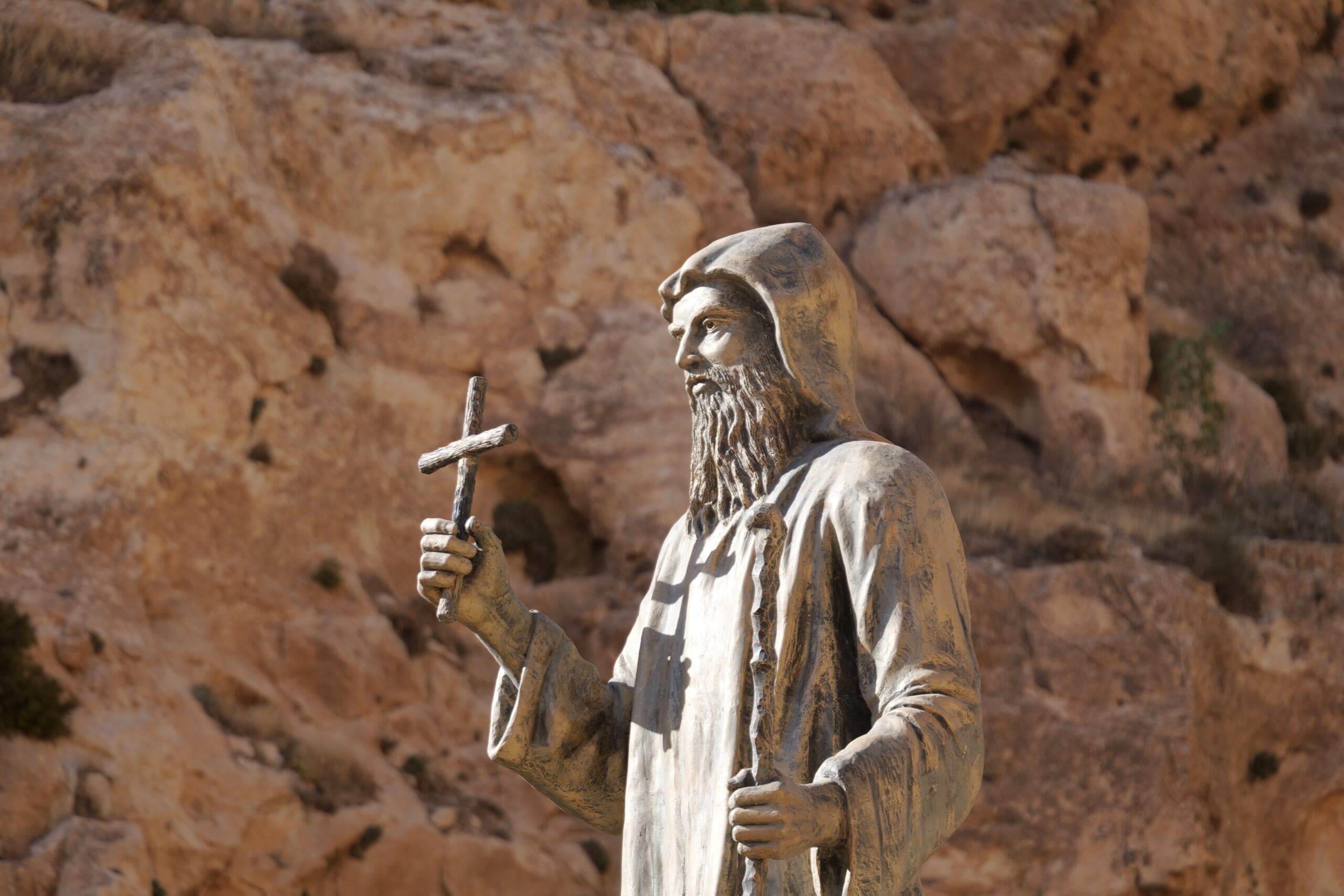Statue of a bearded figure in a hooded robe holding a cross, set against a rocky background, symbolizing faith and spirituality.