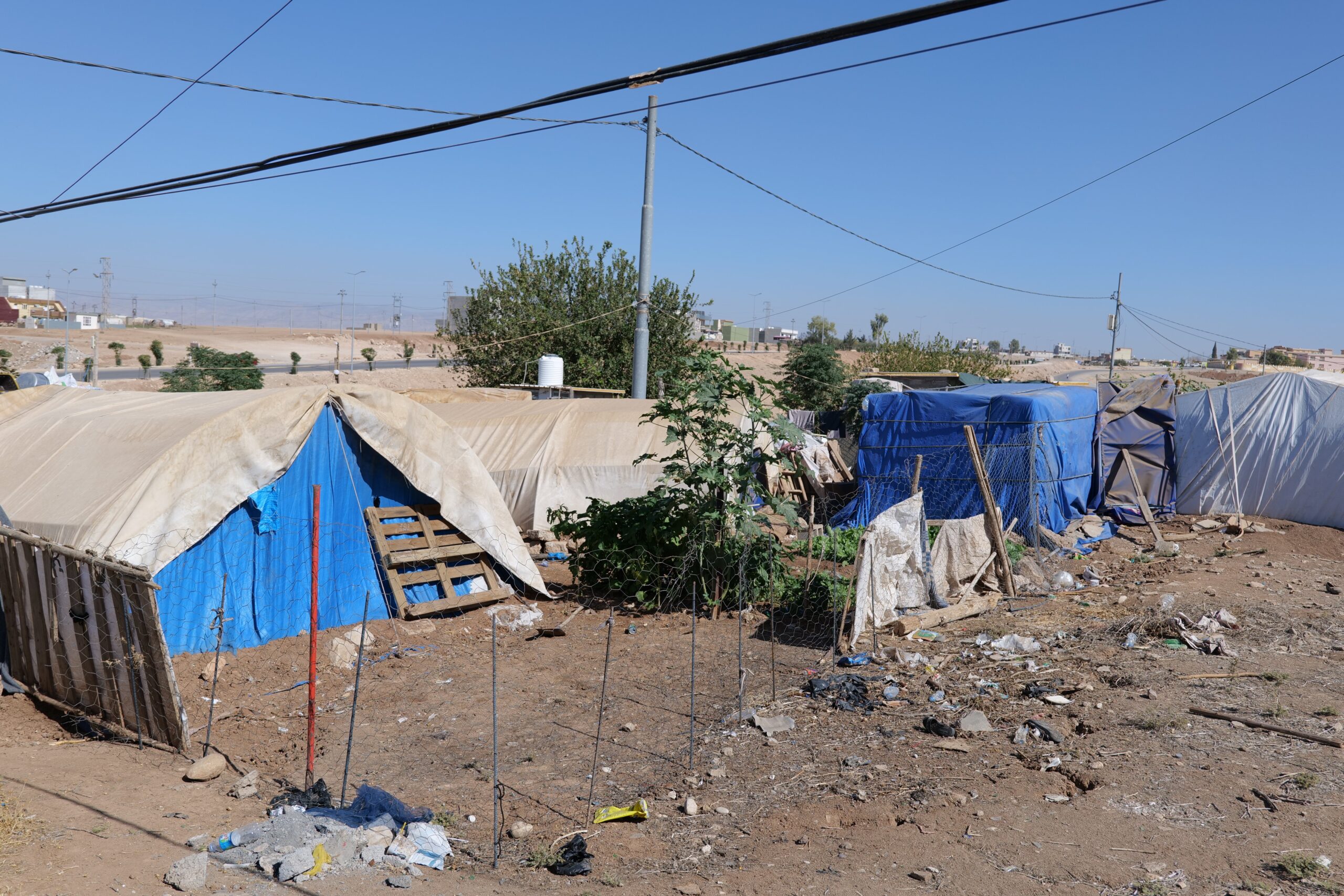 Informal settlement with makeshift tents and debris under a clear blue sky, highlighting living conditions in a vulnerable community.