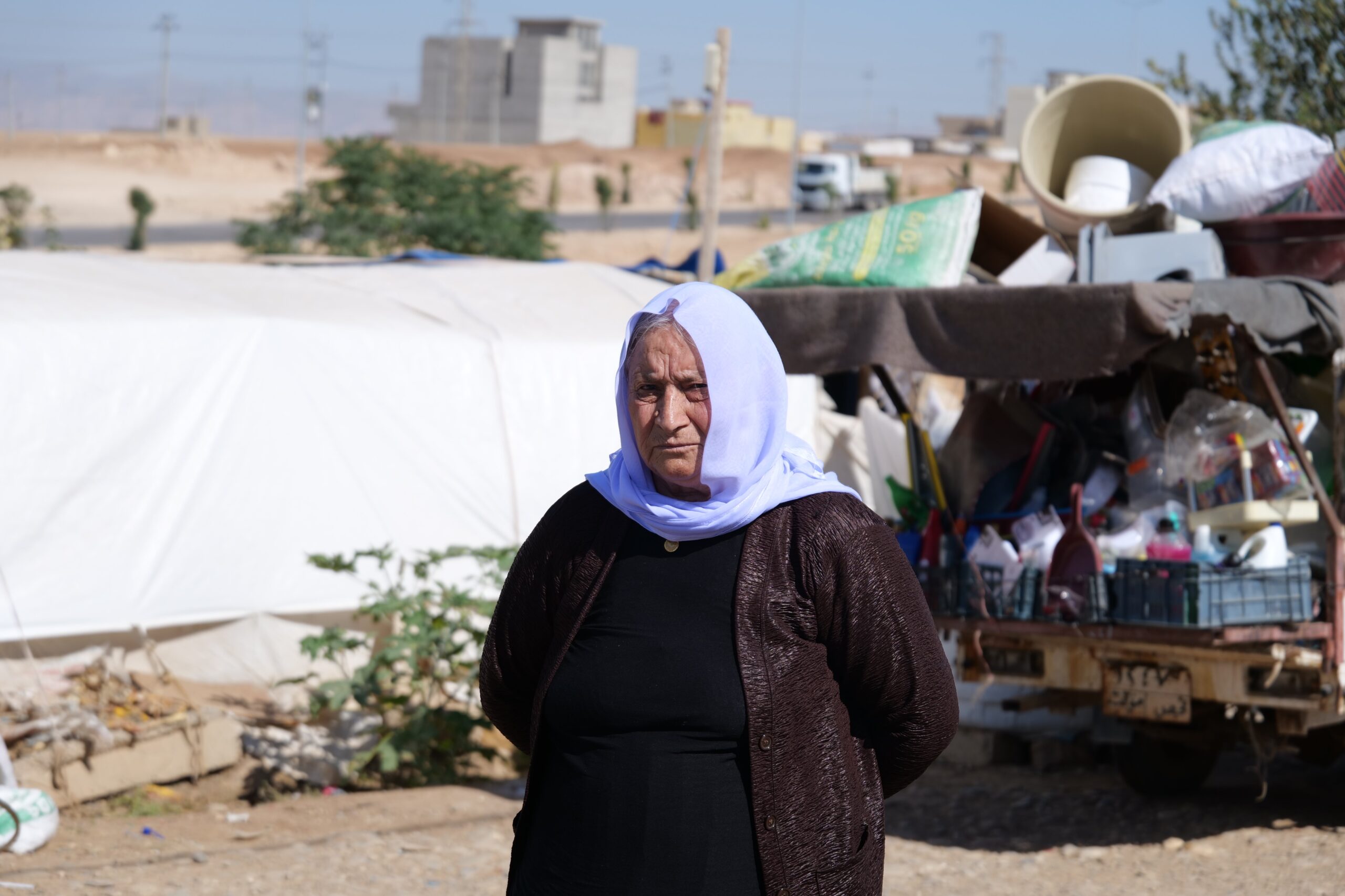 Elderly woman in traditional attire stands in a makeshift settlement surrounded by tents and scattered belongings under a clear blue sky.