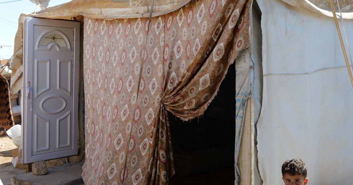 A young boy stands outside a patterned tent with an open door, highlighting living conditions in a refugee camp.