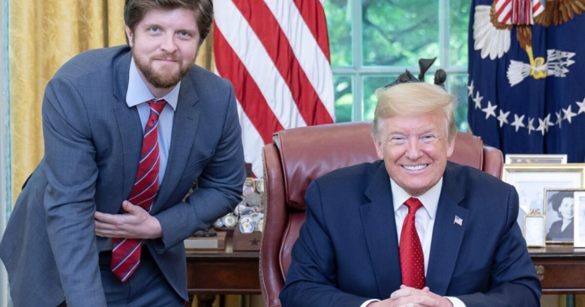 Donald Trump smiles while seated at the Resolute Desk in the Oval Office, accompanied by a man in a suit, with American flags in the background.