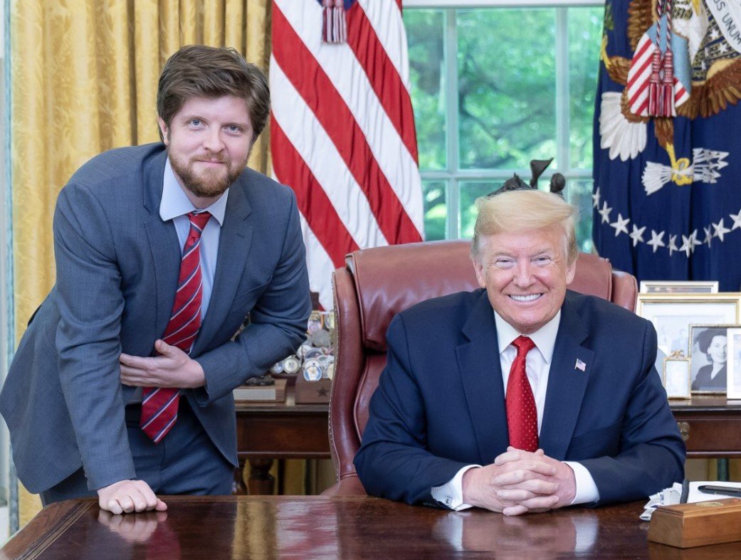 Donald Trump smiles while seated at the Resolute Desk in the Oval Office, accompanied by a man in a suit, with American flags in the background.