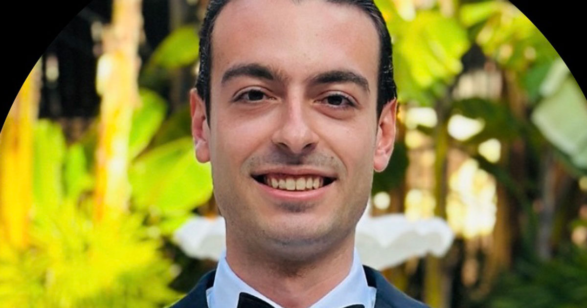 Smiling man in a tuxedo with a bow tie standing outdoors surrounded by lush greenery.