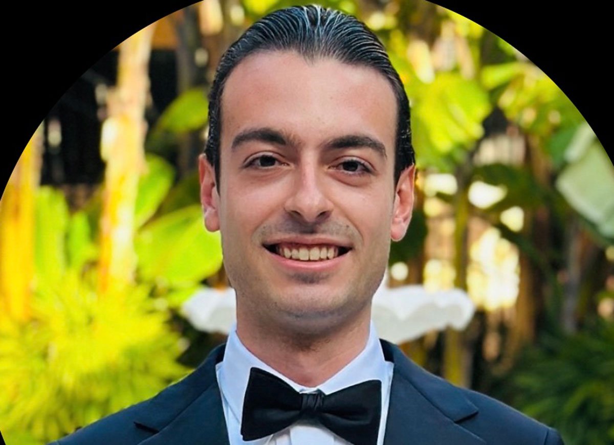 Smiling man in a tuxedo with a bow tie standing outdoors surrounded by lush greenery.