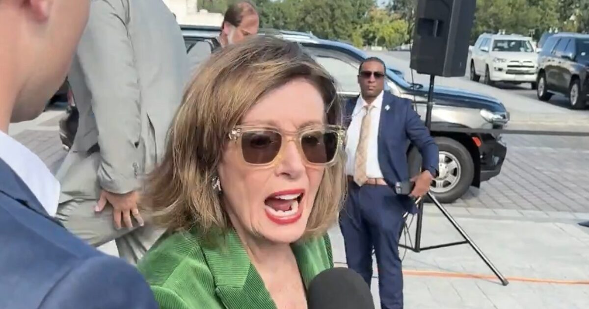 Nancy Pelosi speaking to the media while surrounded by security and vehicles, showcasing her engagement in a public event.